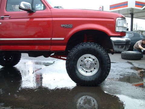 1994 Ford Bronco - A red 1994 Ford Bronco is shown from the front quarter angle, with a person working on a tire in the background.