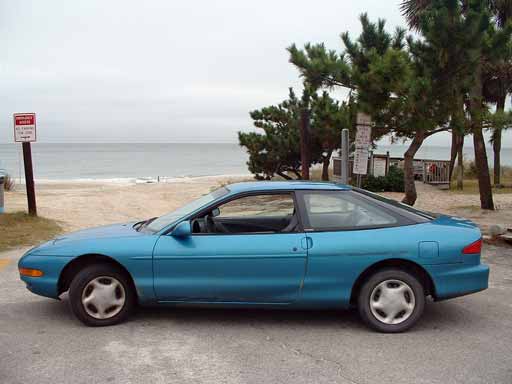 Side view of a blue 1993 Ford Probe parked near a beach with trees and a sign