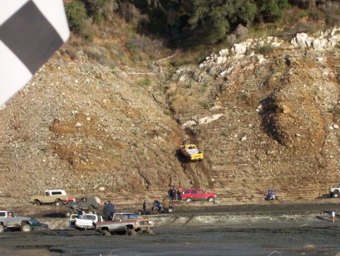 2000 Ford F-450 - Wide shot of an off-road park: a yellow lifted pickup climbs a steep rocky hillside gully while spectators and a red SUV watch from the fla…