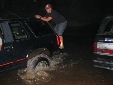 1988 Ford Bronco - Two SUVs are partially submerged in water at night, with one person attempting to get on top of the first vehicle.