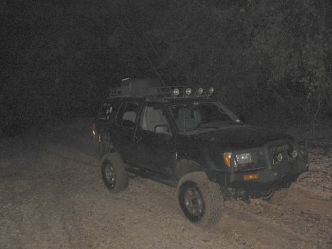 1988 Ford Bronco - A black SUV equipped with a roof rack and multiple auxiliary lights is seen parked on a dirt path during nighttime.
