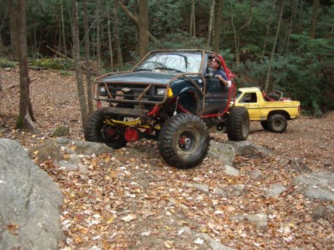 1985 Ford Bronco - A heavily modified dark green SUV navigates rocky terrain, with a yellow pickup truck visible in the background.