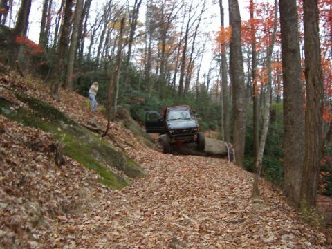 1985 Ford Bronco - A dark lifted 4x4 with its driver's door open descends a steep, leaf-covered woodland trail, front wheels dropping over a rock ledge. A per…