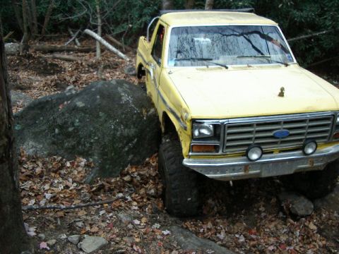 1985 Ford Bronco - Three-quarter front view of a yellow lifted Ford Bronco with black hardtop flexing on a leaf-covered trail beside a large mossy boulder, fr…