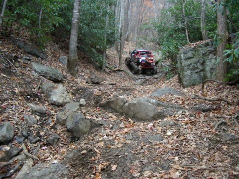 1985 Ford Bronco - A red off-road vehicle navigates a challenging, rocky path through a dense forest.