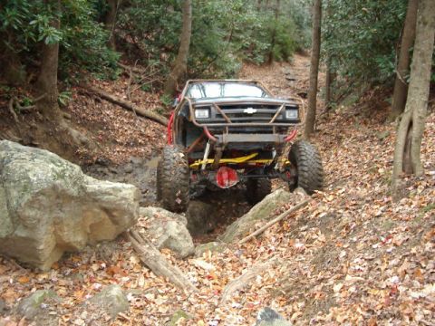1985 Ford Bronco - This heavily modified off-road vehicle navigates a challenging, muddy, and rocky trail through a wooded area.