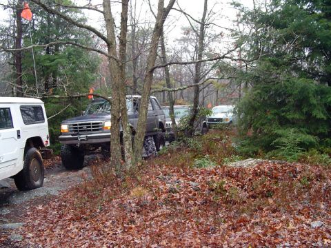 1979 Ford Bronco - Several off-road vehicles navigate a wooded path covered in fallen leaves.