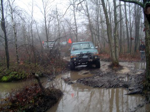 1979 Ford Bronco - Multiple SUVs traverse a wet and muddy path through a dense forest on an overcast day.