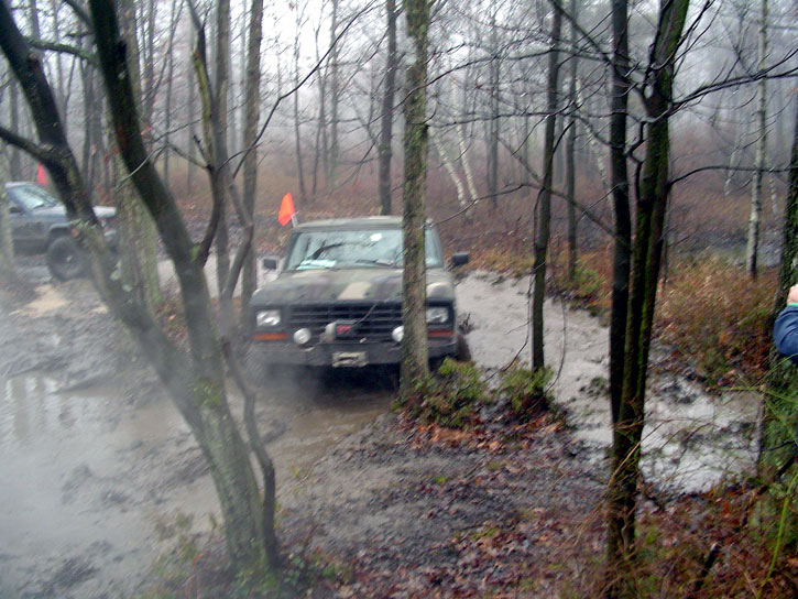 Two off-road vehicles navigating a muddy trail through woods; one in foreground has winch