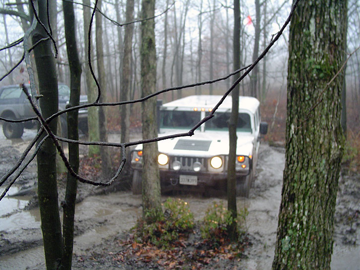 White Hummer H2 and a dark SUV driving off-road in a muddy forest