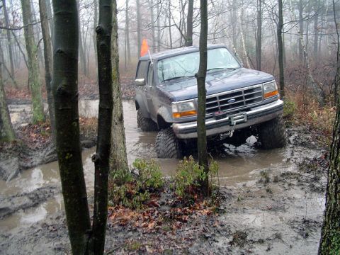 1979 Ford Bronco - A Ford Bronco navigates a muddy trail in a forest, with large tires splashing through the water.