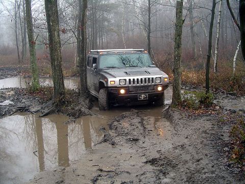 1979 Ford Bronco - A Hummer H2 tackles a challenging muddy trail surrounded by trees.