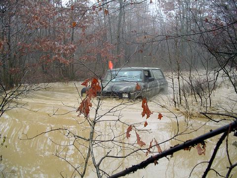 1979 Ford Bronco - A dark SUV navigates through a flooded wooded area, with water reaching its doors.