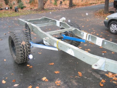1978 Ford Bronco - A bare truck frame in light gray primer sitting on a leaf-covered driveway during a suspension mock-up, with blue-painted radius arms and a…