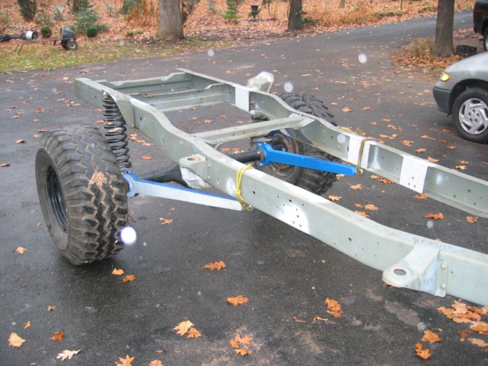 Bare primered truck frame with blue radius arms and coil spring test-fitted on a driveway