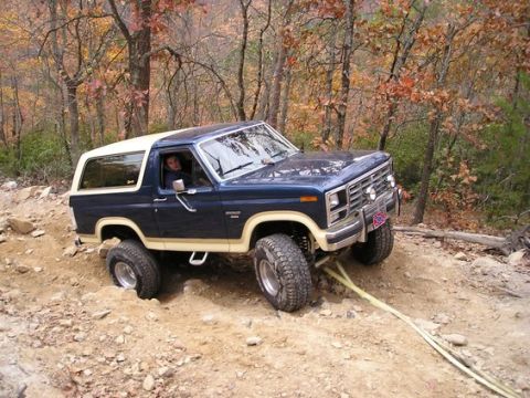 1985 Ford Bronco - A blue and tan Ford Bronco navigates a challenging off-road trail with its front right tire stuck.