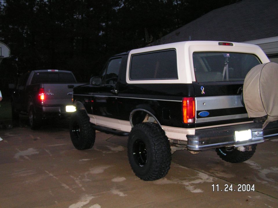 Lifted black and tan Ford Bronco with big mud tires, rear three-quarter view in a driveway at dusk
