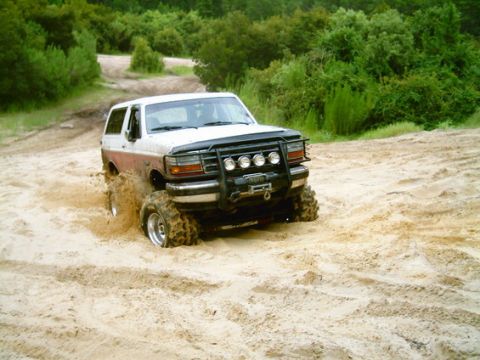 1993 Ford Bronco - A white and brown Ford Bronco navigates sandy terrain, its front tires partially submerged in the sand.