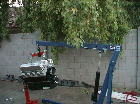 1979 Ford Bronco - An engine is suspended by a blue engine hoist in front of a cinder block wall.