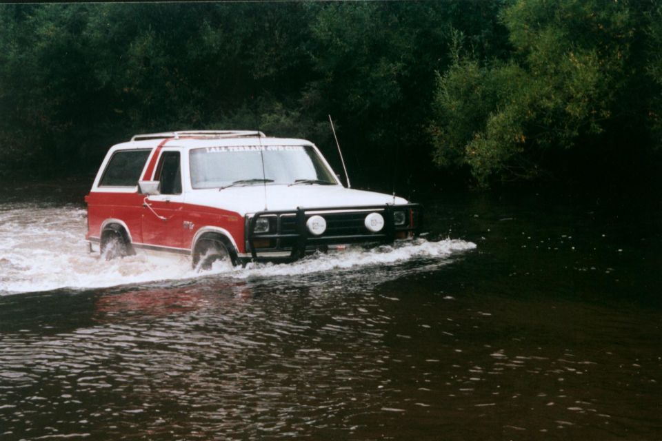 Red and white Ford Bronco crossing a river with water at bumper height, bull bar and driving lights visible
