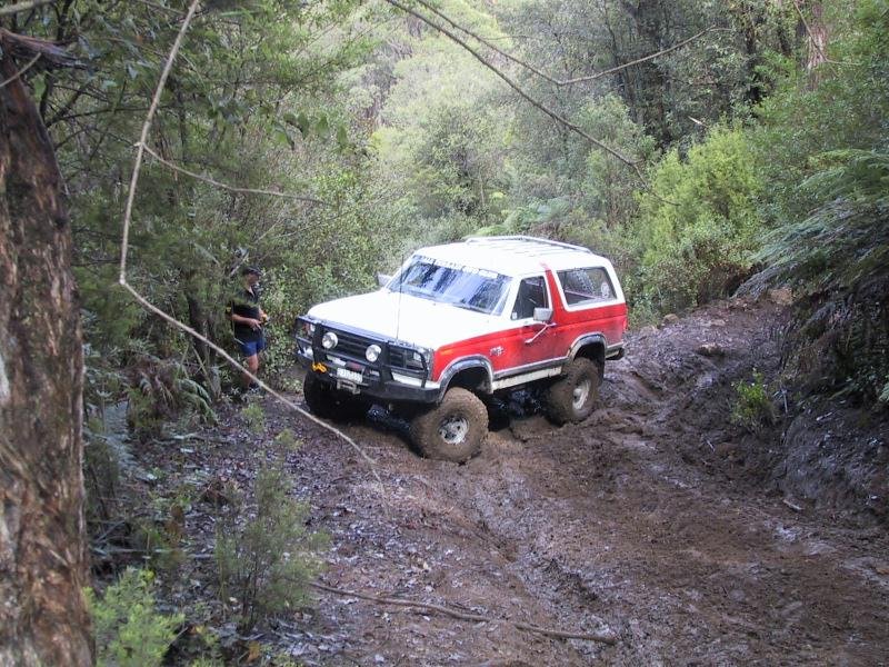 White and red lifted Ford Bronco on muddy off-road forest trail with spotter nearby