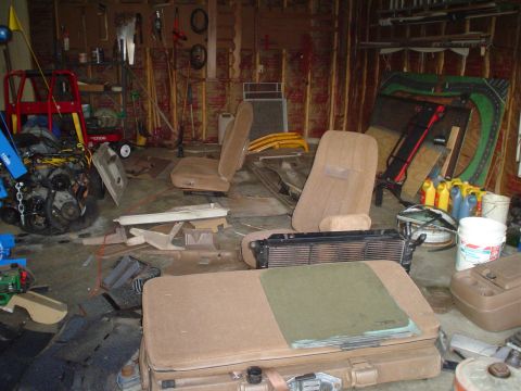 1981 Ford Bronco - A cluttered garage contains car seats, an engine, a radiator, and other automotive parts.