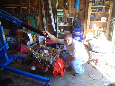 1981 Ford Bronco - A man crouches next to an engine being lifted by a blue engine hoist in a cluttered garage.
