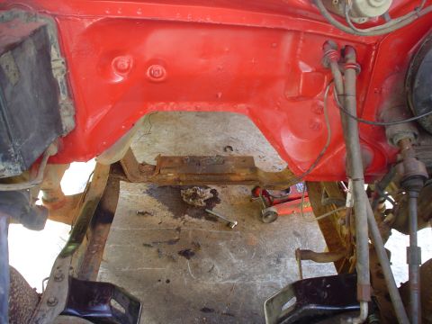 1981 Ford Bronco - Close-up view of the red painted engine bay frame and rusty crossmember, showing some of the vehicle's undercarriage.