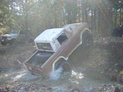 1974 Ford Bronco - A 1974 Ford Bronco navigates a water obstacle in a wooded area, with another vehicle visible in the background.