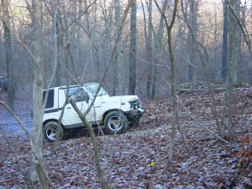 White Suzuki Samurai driving on a wooded trail with fallen leaves