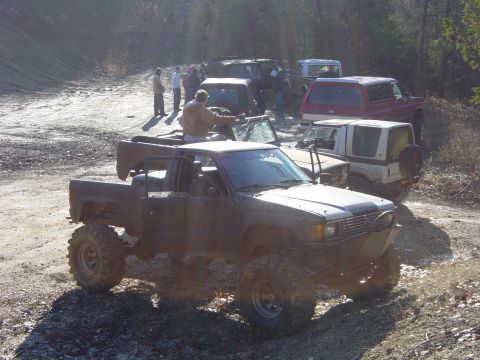 1974 Ford Bronco - A group of off-road vehicles are gathered on a dirt path, surrounded by trees.