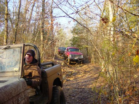 1974 Ford Bronco - A group of off-road vehicles, including a tan 1974 Ford Bronco, navigate a challenging rocky trail through a wooded area.