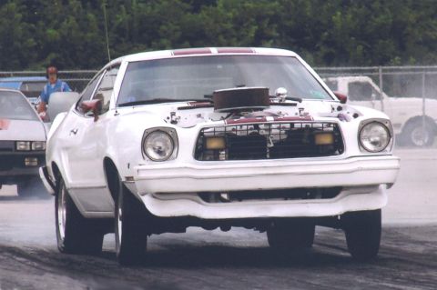1976 Ford Mustang II - A white 1976 Ford Mustang II with racing stripes and an exposed engine sits on a drag strip.