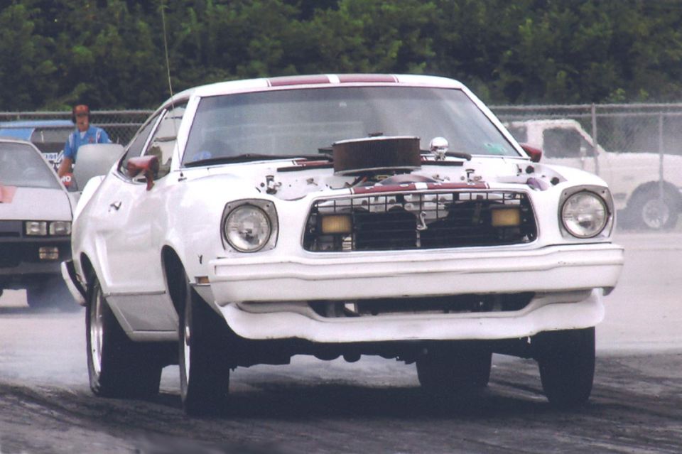 White 1976 Ford Mustang II with hood open, showing engine and air intake, on a race track