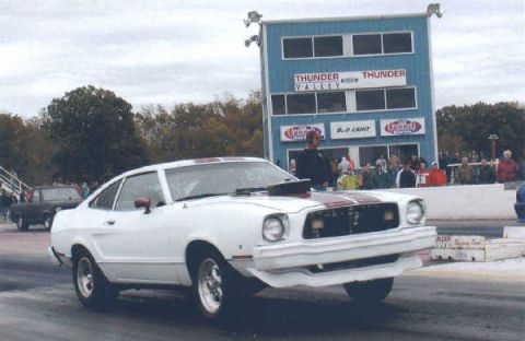 1976 Ford Mustang II - White 1976 Ford Mustang II with red stripes on a drag strip, with a spectator stand in the background.