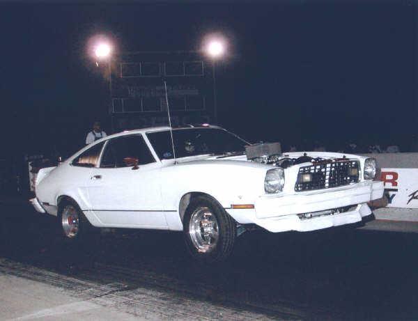 White drag car with hood scoop, front wheels off the ground at a drag strip