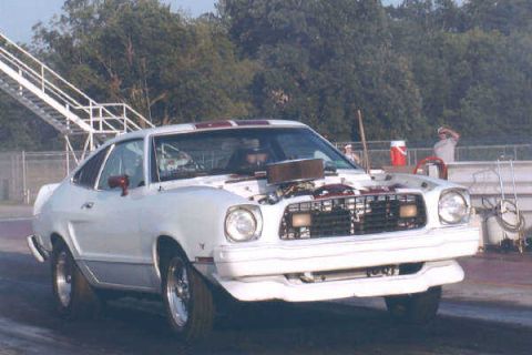 1976 Ford Mustang II - A white coupe with racing stripes and an open hood is on a track, possibly at a drag strip.