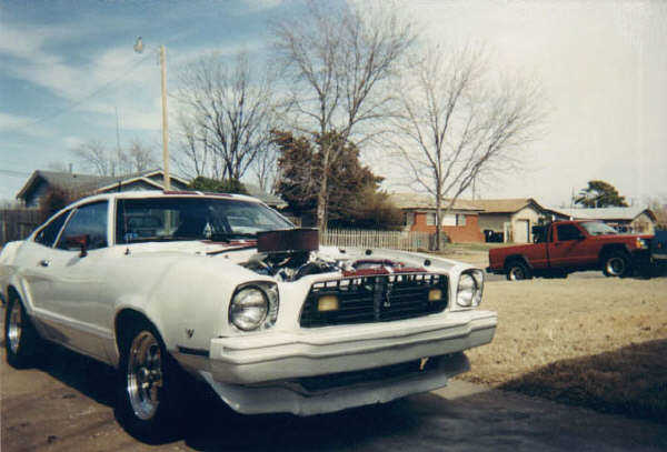 White 1976 Ford Mustang II with hood open, showing engine. Red pickup truck in background