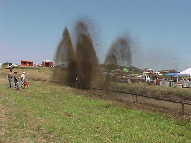 Off-road vehicles creating large mud spray at an event with spectators and tents in the background
