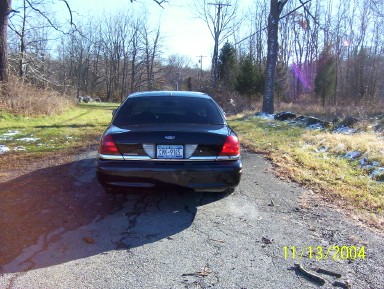 1999 Ford Crown Victoria - A black 1999 Ford Crown Victoria is parked on a dirt road with trees and snow in the background.