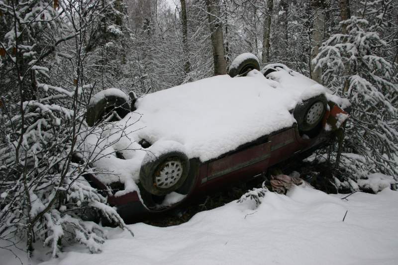 Overturned red car covered in snow, resting on its side in a wooded area