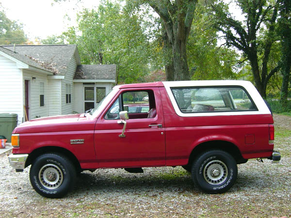 Red two-door SUV with white hardtop, side view, parked on gravel by a white house.