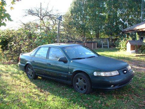 1993 Ford Taurus - A green 1993 Ford Taurus sedan is parked on a grassy lawn in front of trees and a small building.