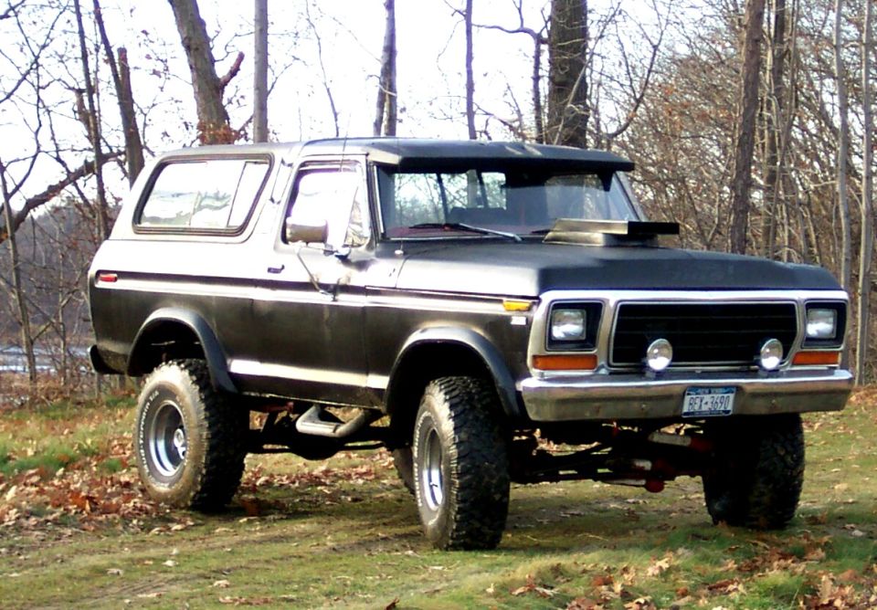 Lifted black-and-silver full-size Ford Bronco on mud tires parked on grass among bare trees