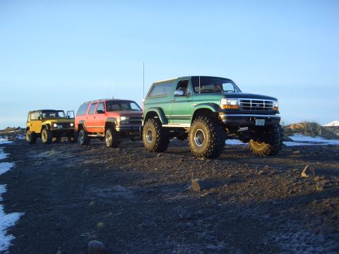 1996 Ford Bronco - Four off-road vehicles are lined up on a dirt terrain under a clear blue sky.