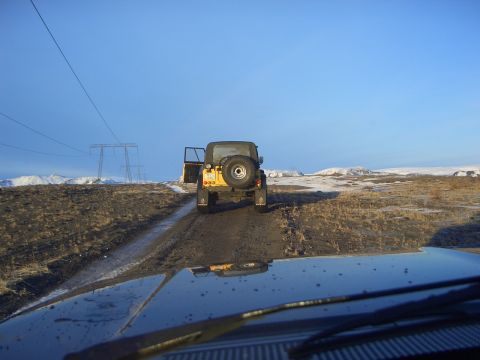 1996 Ford Bronco - A yellow Jeep Wrangler travels down a dirt road, with another vehicle's hood visible in the foreground and a vast, snowy landscape behind.
