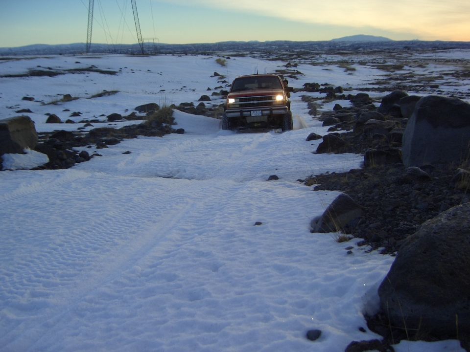 Red Ford Bronco with headlights on driving on snowy trail at dusk