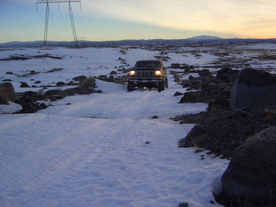 Dark Ford Bronco with headlights on driving on a snowy rocky trail at dusk