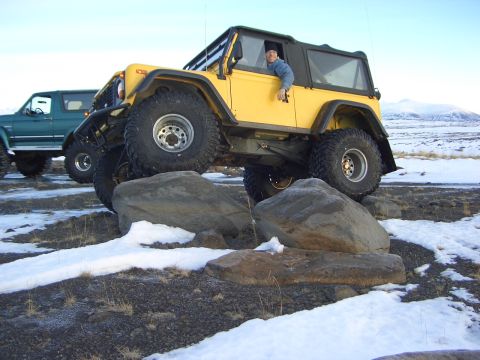 1996 Ford Bronco - A person drives a modified yellow Jeep over large rocks in a snowy landscape, with another vehicle visible behind.