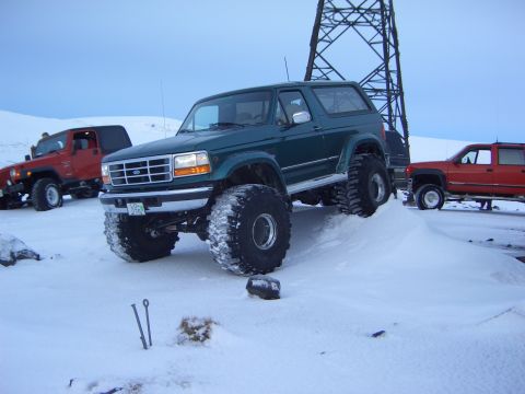 1996 Ford Bronco - A green Ford Bronco with oversized tires is parked on a snow-covered hill next to other vehicles.